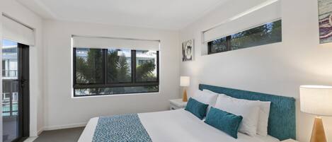 Une chambre moderne avec un grand lit, une fenêtre donnant sur des palmiers, une table de chevet avec une lampe et un luminaire au plafond.