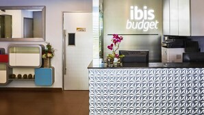 Un área de recepción con diseño moderno, que incluye un mostrador con patrón, un letrero que dice 'ibis budget' y un espejo en la pared.