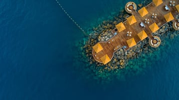 An aerial view of a wooden deck with tables and umbrellas, surrounded by clear water and rocks.