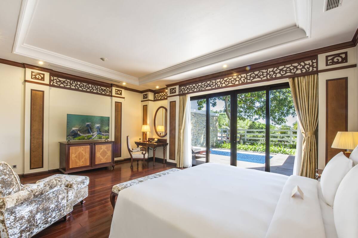 Een hotelkamer met een groot bed, een bureau met stoel, een televisie en uitzicht op een buitenzwembad.
