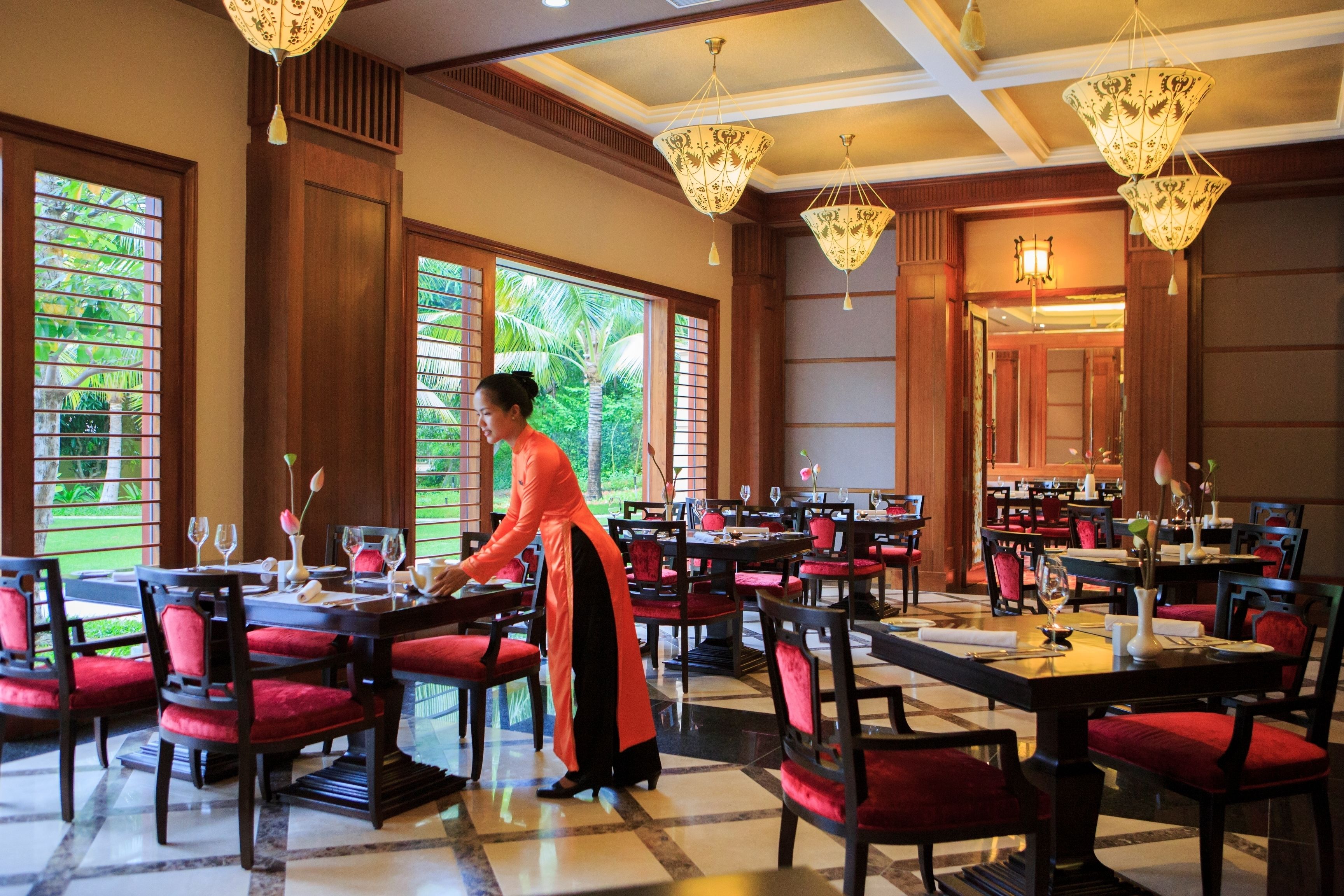 A dining area with tables set for service, a waiter in traditional attire, and large windows with a view of greenery.