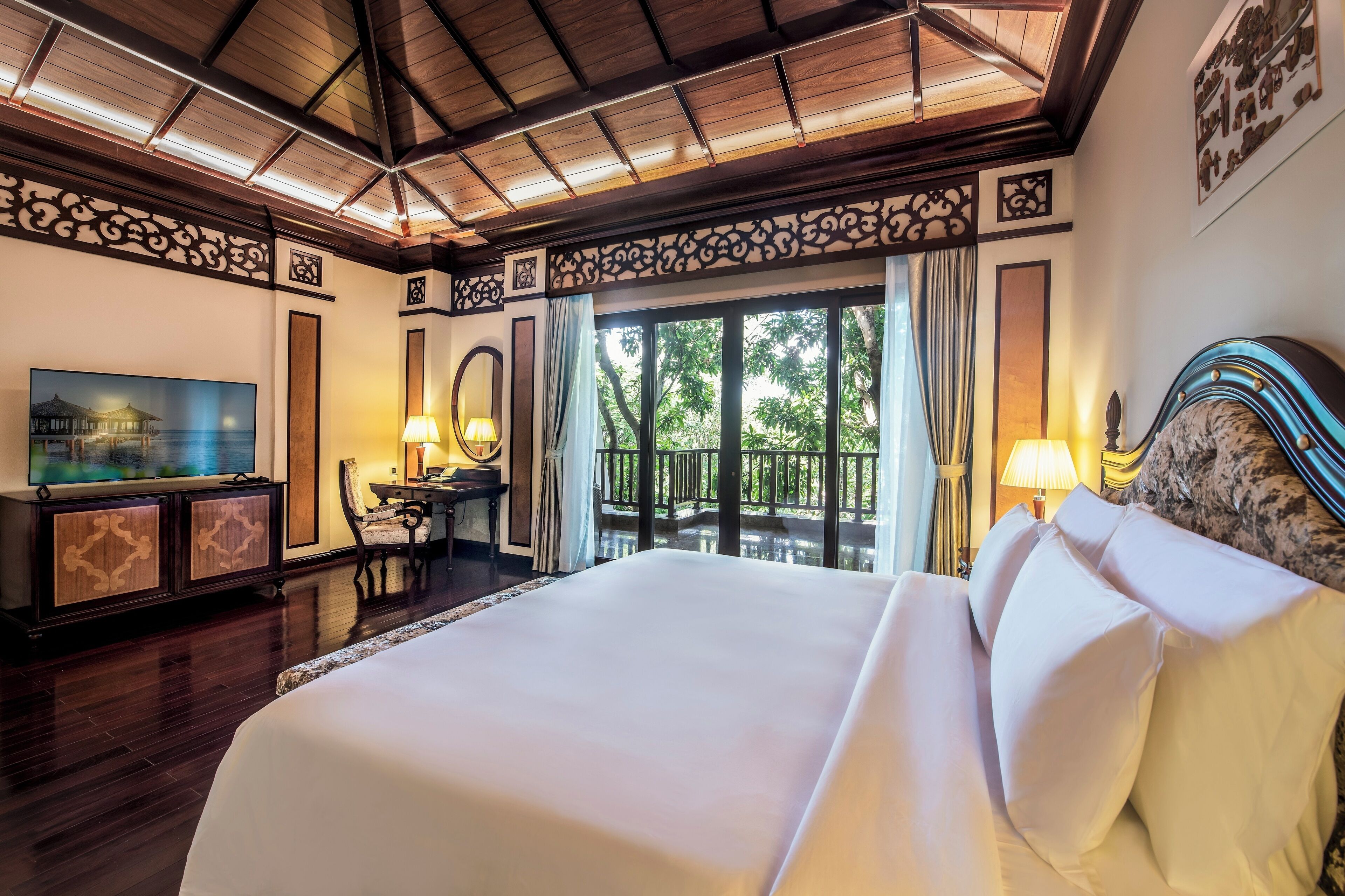 Habitación de hotel con una cama grande, un escritorio con lámpara, televisor y balcón con vistas a árboles.