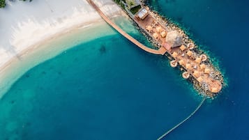 Ein Luftbild eines Resorts mit einer Holzsteg, der sich in türkisfarbenes Wasser erstreckt, umgeben von weißem Sand und klarem, blauem Meer.
