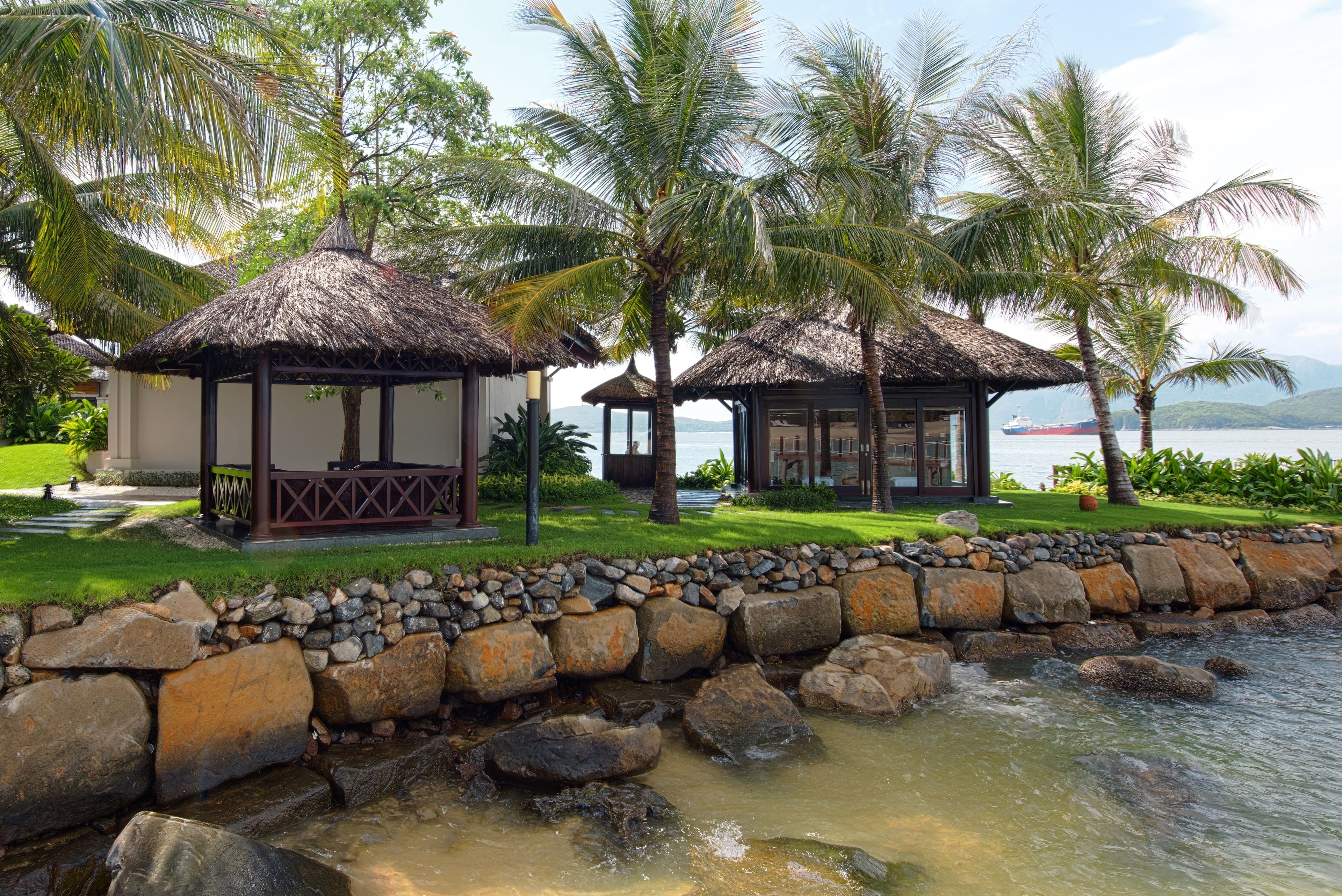 A tranquil resort setting with thatched-roof structures, a stone retaining wall, and a calm water body.