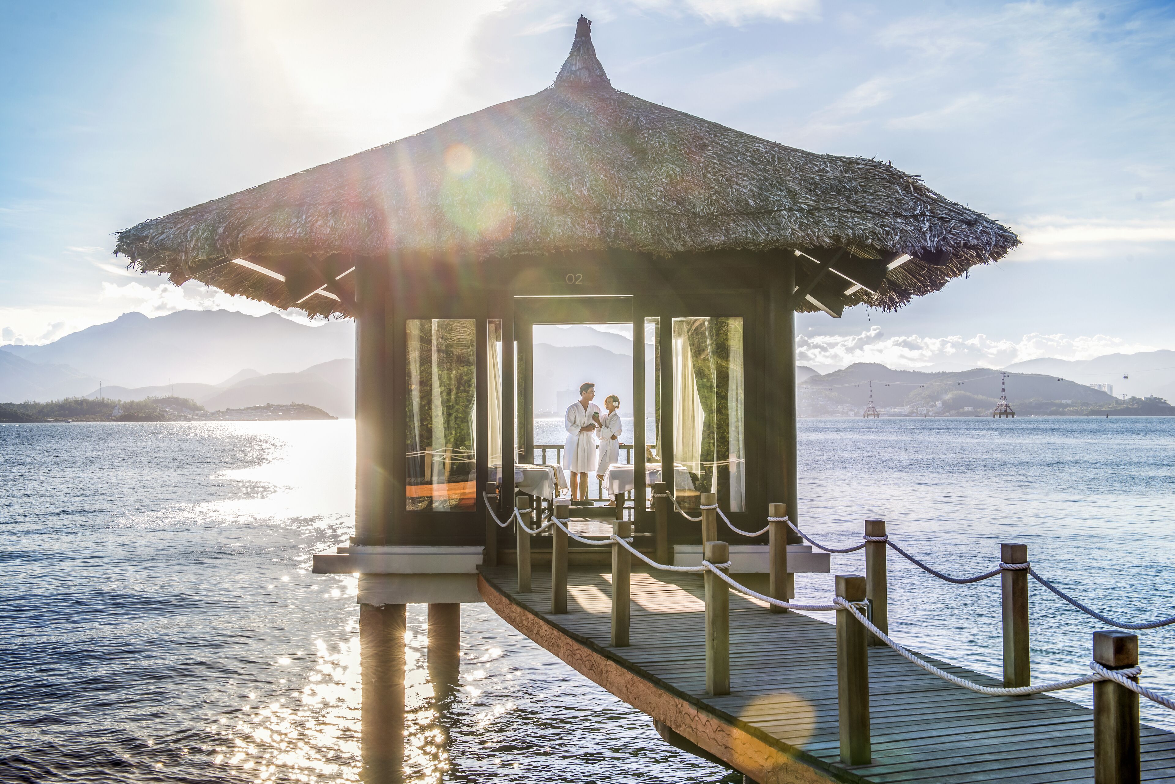 A wooden pier leading to a thatched-roof gazebo with a view of the sea and mountains in the background.