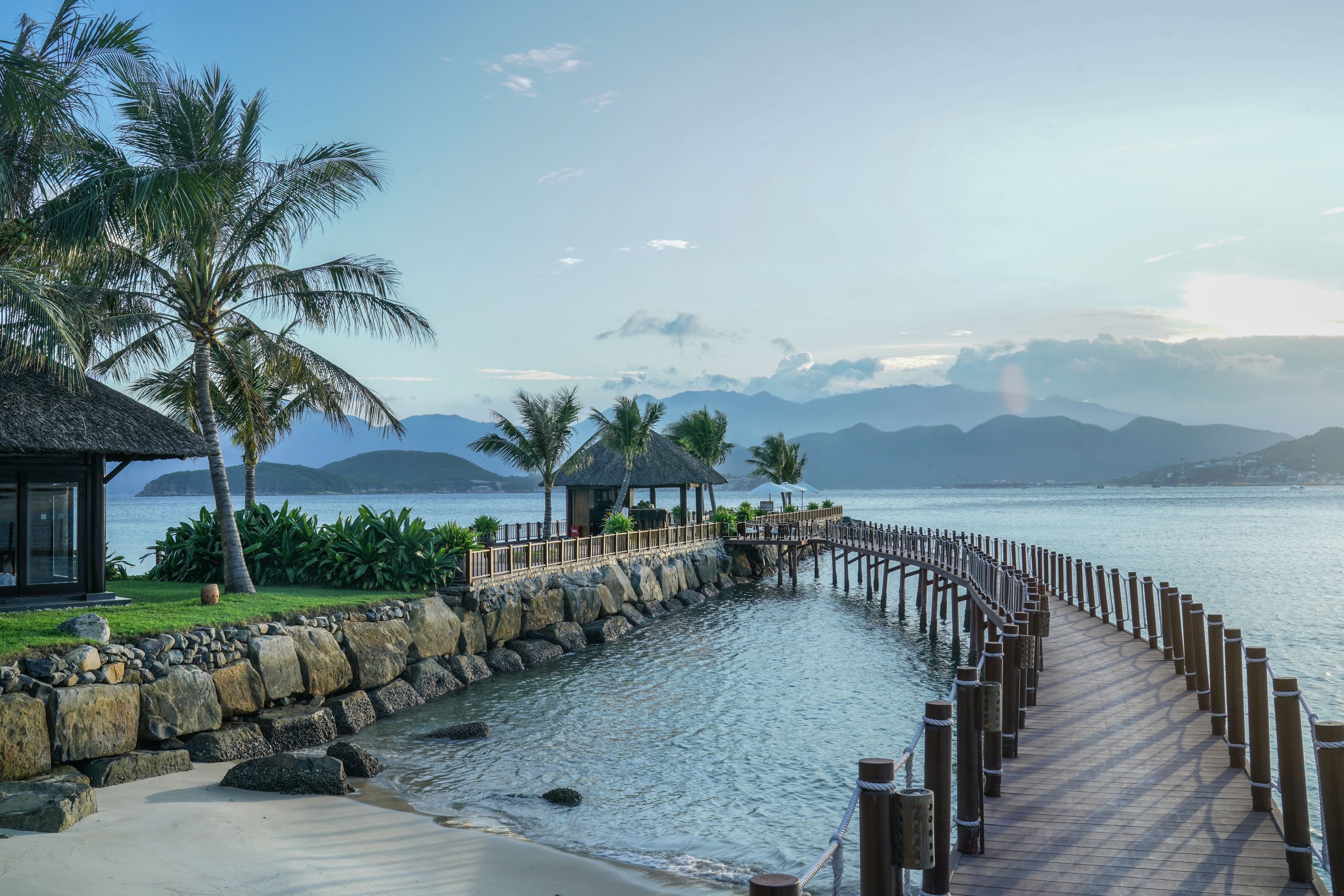 A wooden boardwalk leading to a beach with palm trees and a thatched-roof structure.
