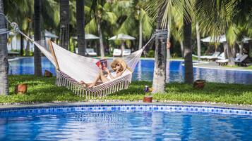 Relax in a hammock between palm trees, surrounded by lush greenery and a clear pool.
