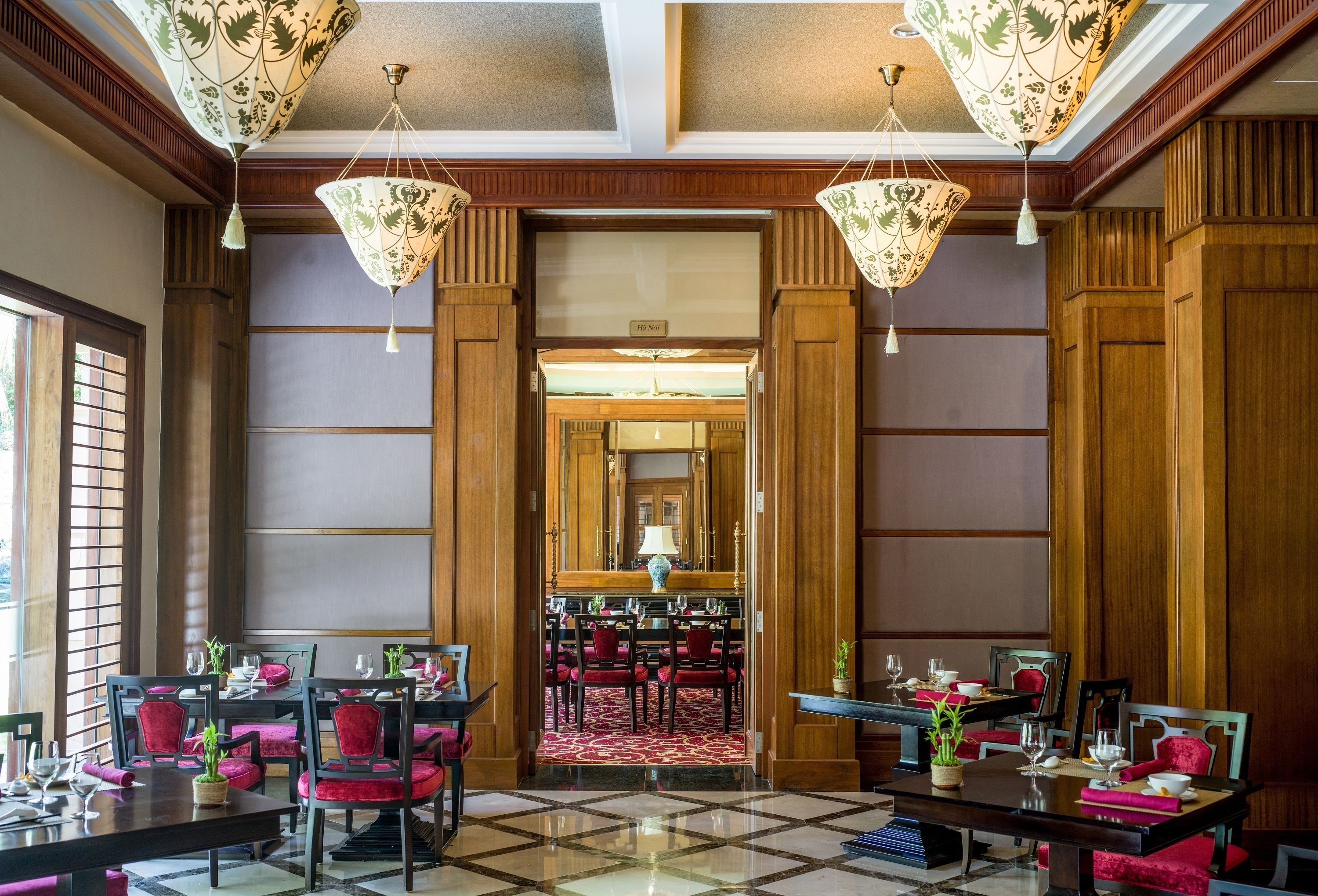 Un vestíbulo de hotel con suelo de cuadros, paneles de madera y sillones tapizados en rojo. Se observan mesas dispuestas para comer con cristalería y una lámpara.