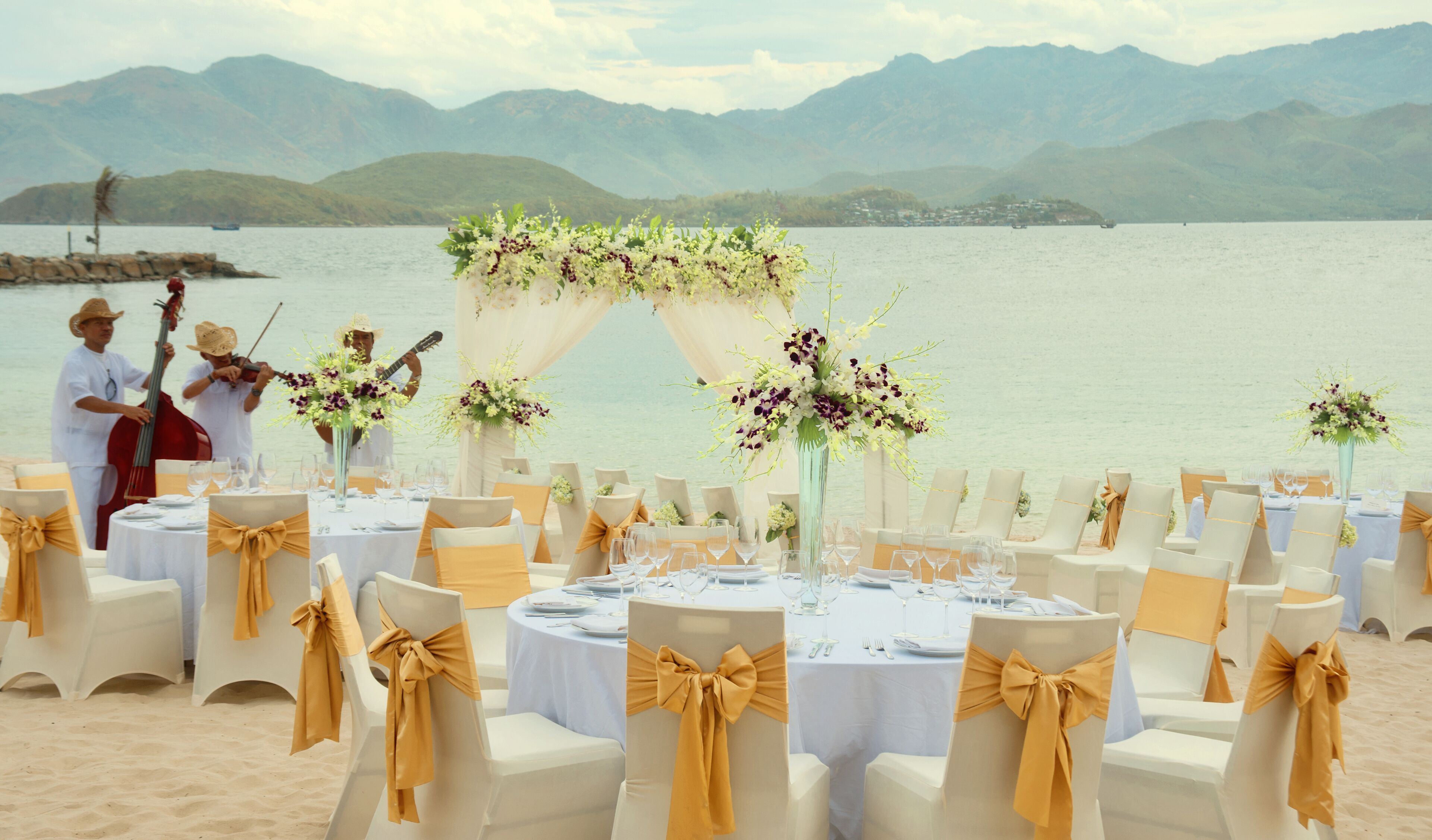A beachside wedding setup with round tables, chairs, and floral centerpieces.