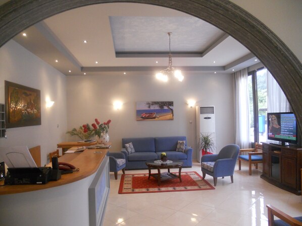 A spacious living area with a blue sofa, a coffee table, and a TV. There is a wooden reception desk with a phone and a computer.