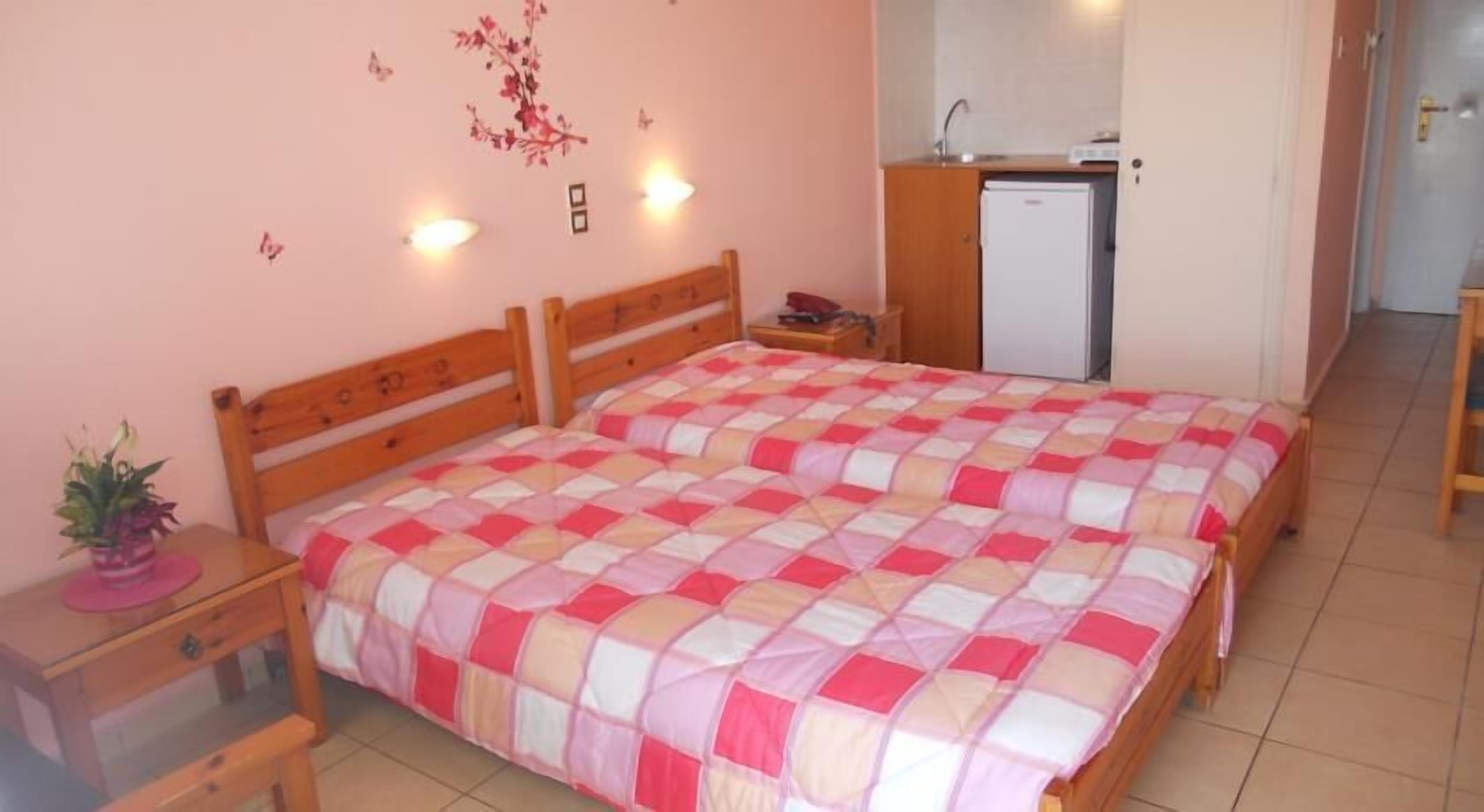 A bedroom with a wooden bed, a bedside table, a small desk, a refrigerator, and a wall-mounted light.