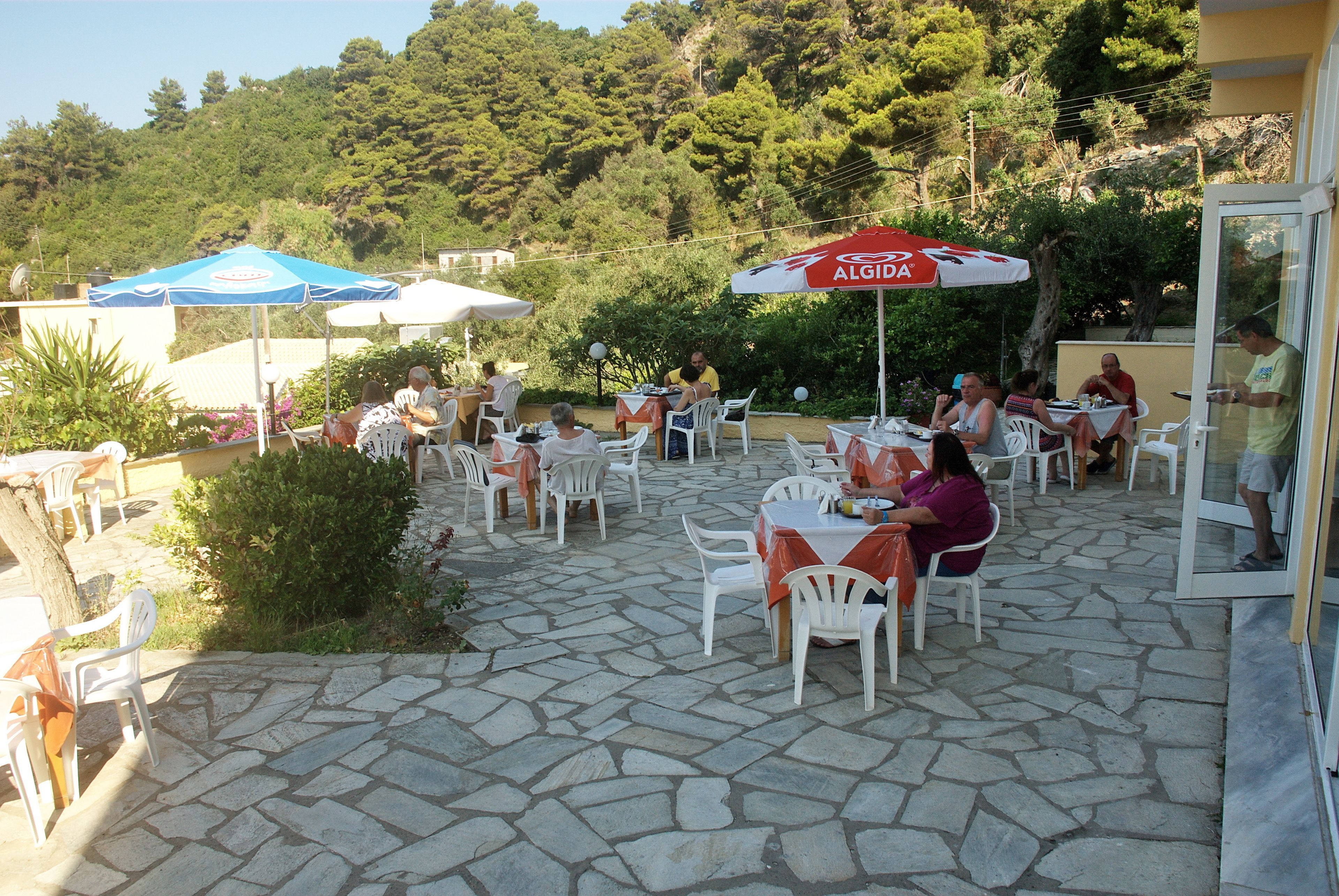 Un patio al aire libre con suelo de piedra, sillas de plástico blanco y mesas bajo sombrillas.