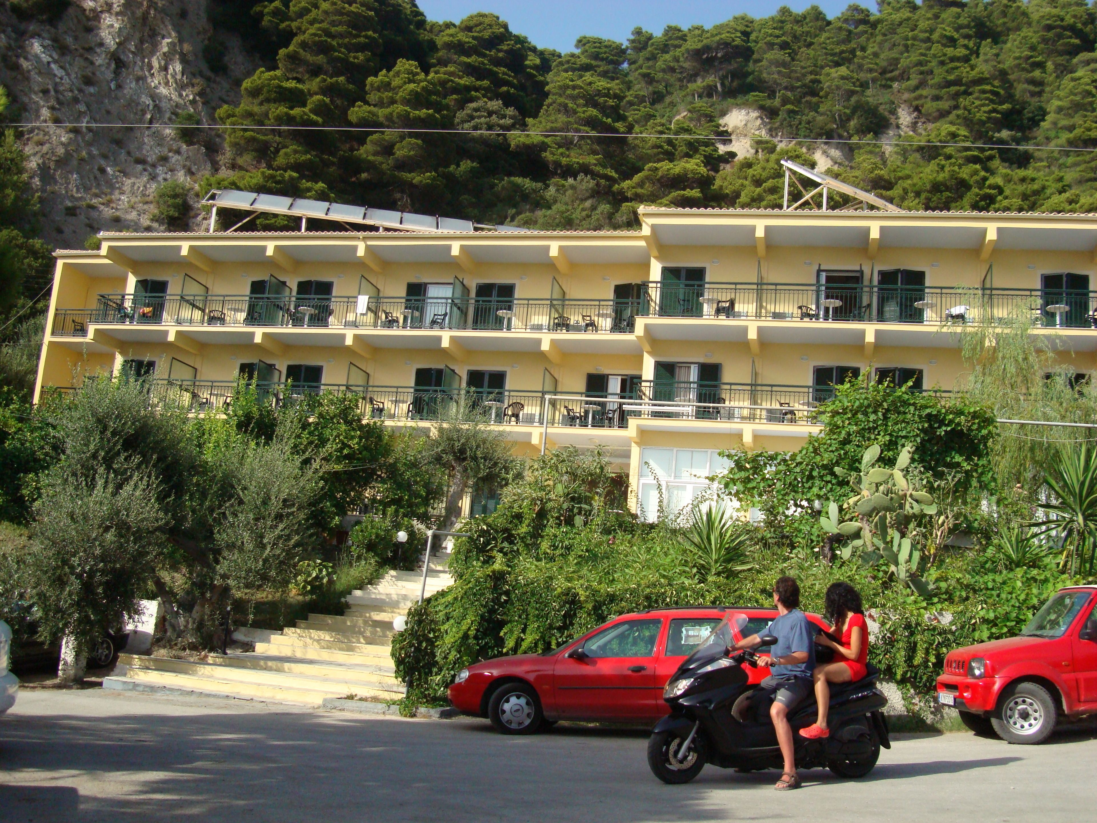 Un edificio amarillo de varios pisos con balcones, un coche rojo y una motocicleta con pasajeros.