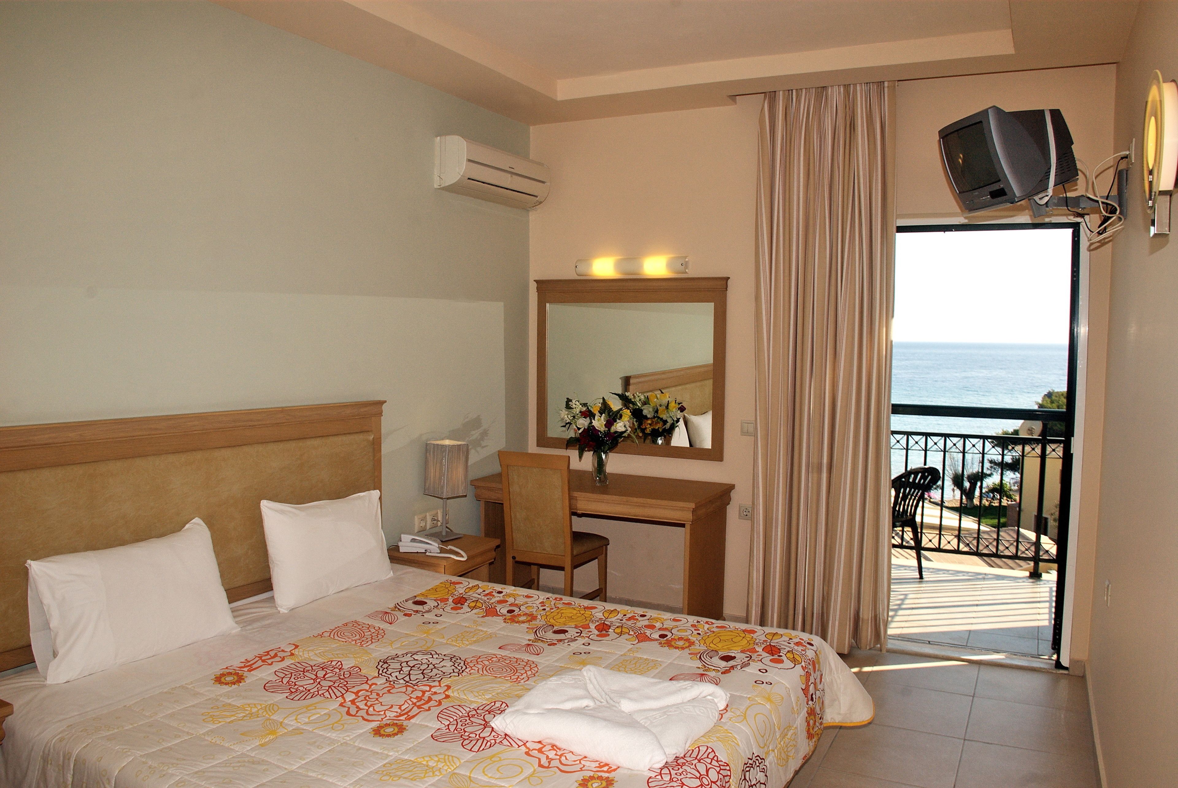 A hotel room with a bed, a desk, a chair, a TV, and a balcony with a view of the sea.