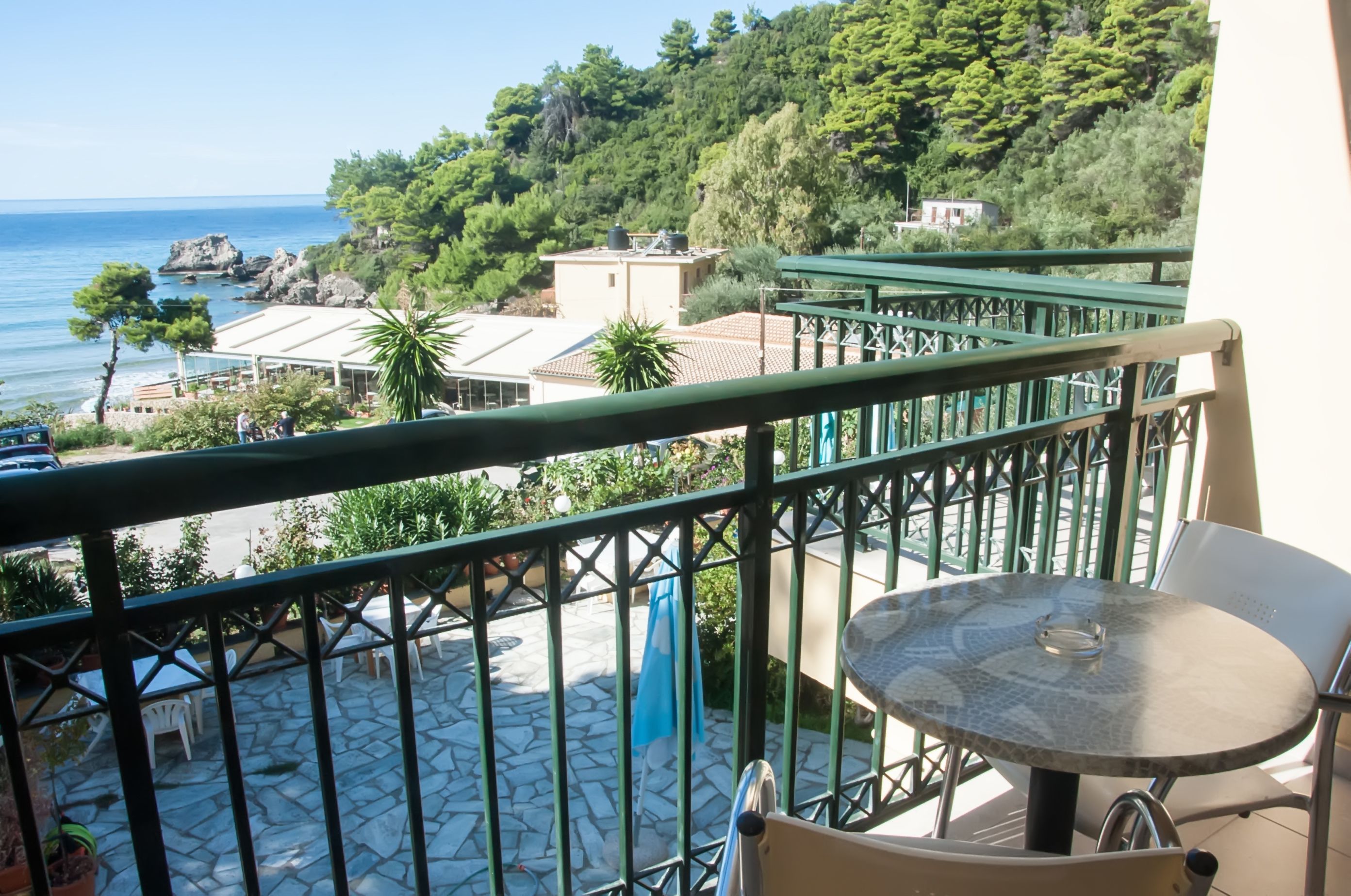 A balcony with a stone floor, a table, and chairs, overlooking a coastal area with trees and buildings.