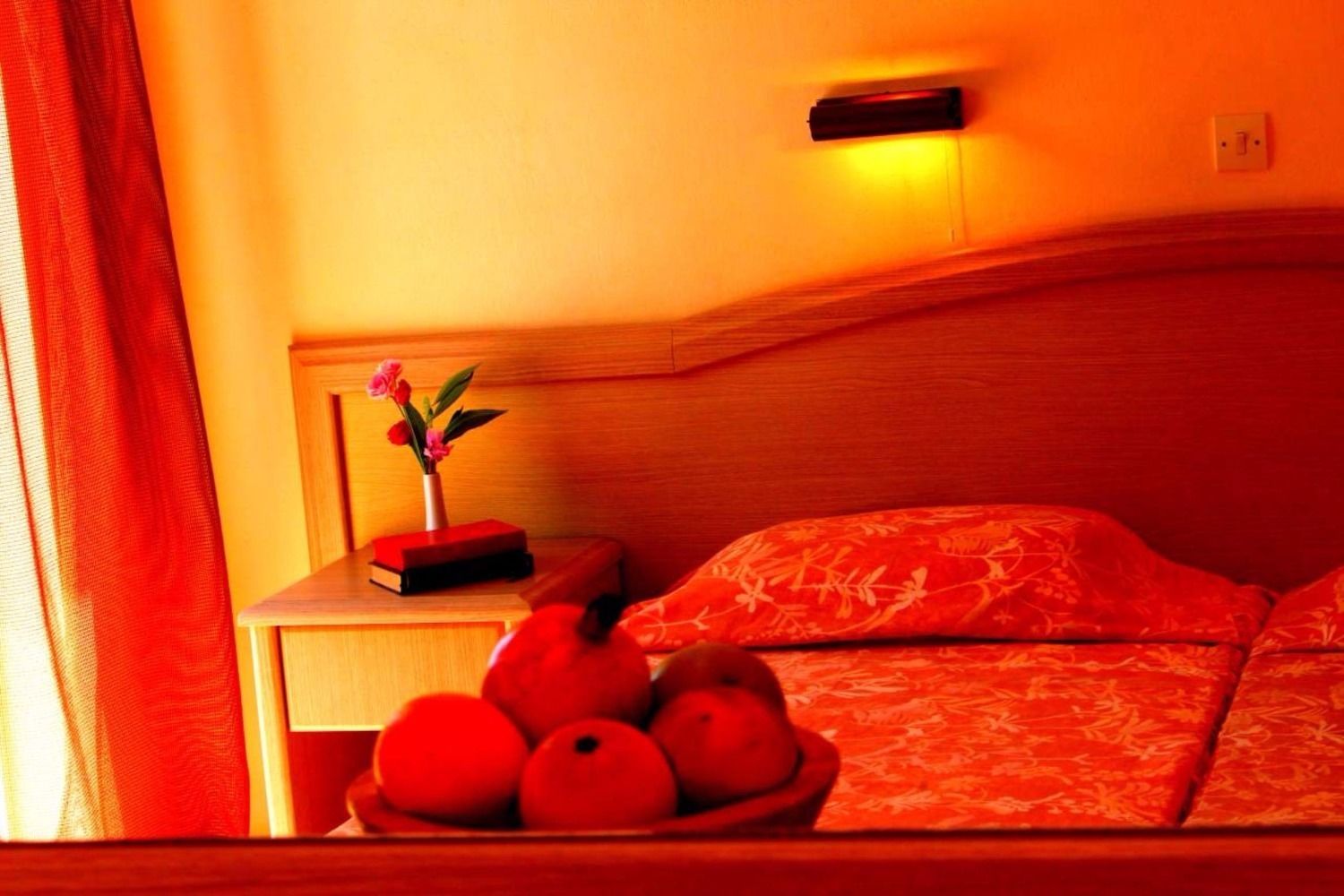 A room with a bed, a bedside table with a vase of flowers, and a bowl of oranges.
