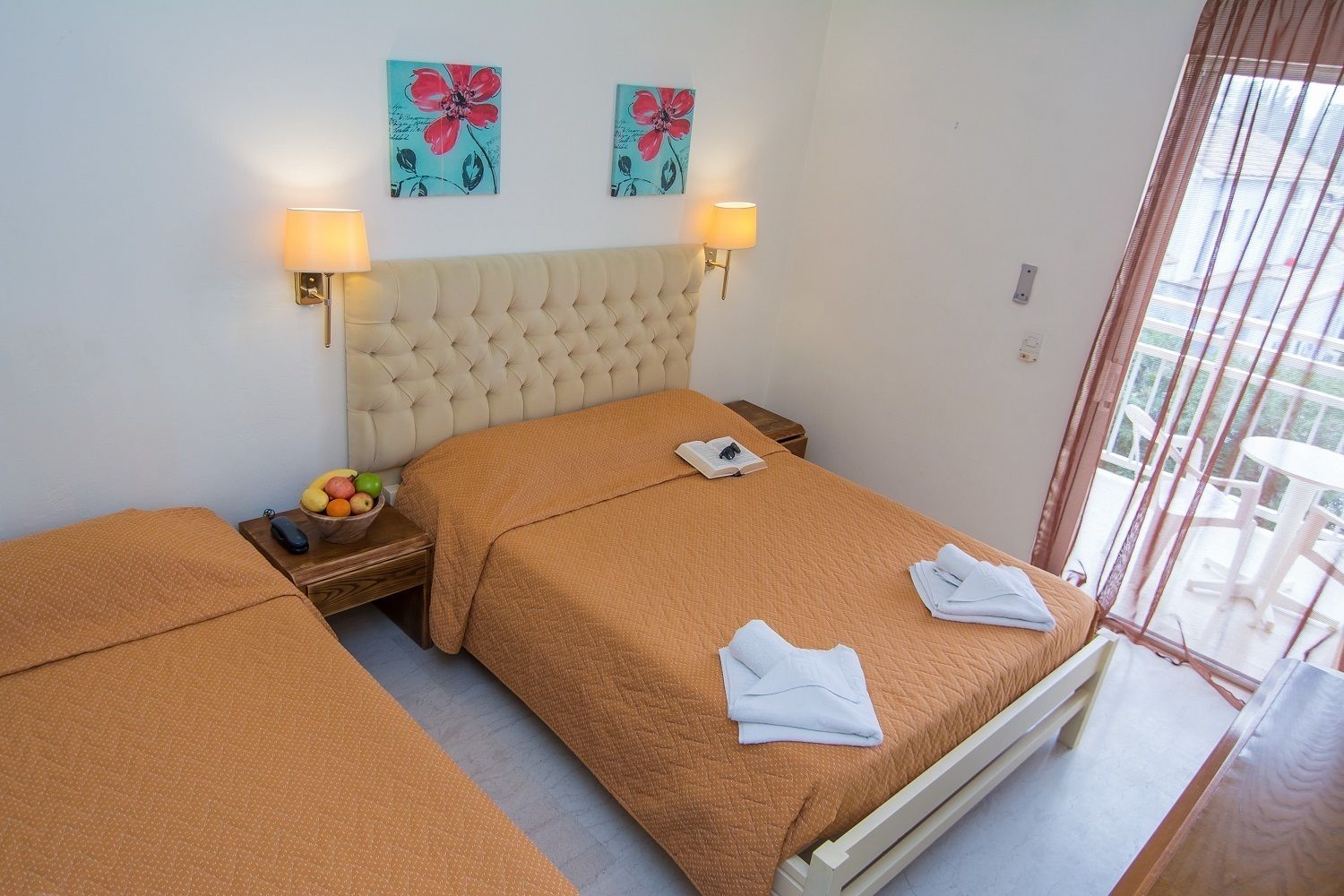 A hotel room with two beds, a fruit bowl, a bedside table, and artwork on the wall.