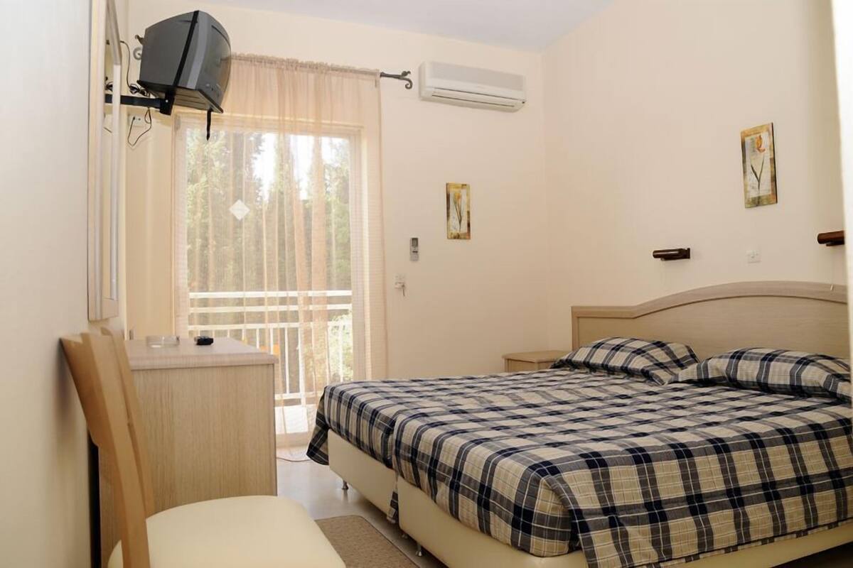 A bedroom with a bed, a desk, and a chair. There is a window with curtains and an air conditioning unit on the wall.