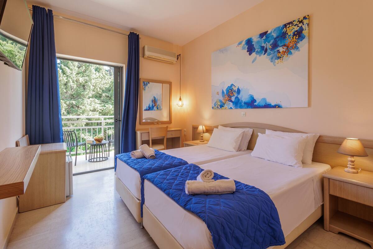 A hotel room with two beds, a desk, a chair, a balcony with a view, and a painting on the wall.