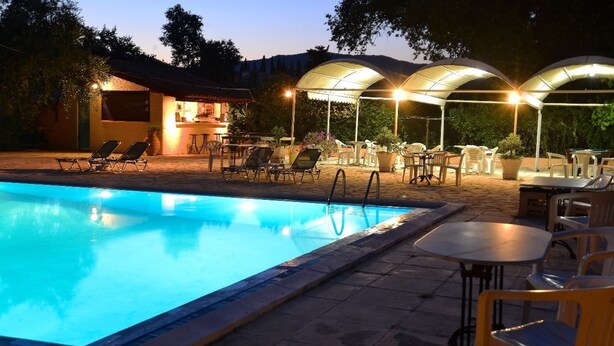 A swimming pool with lounge chairs and tables, surrounded by covered seating areas and a building with a tiled roof. - Primavera Hotel (Corfu)
