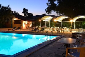 A swimming pool with lounge chairs and tables, surrounded by covered seating areas and a building with a tiled roof. - Primavera Hotel (Corfu)