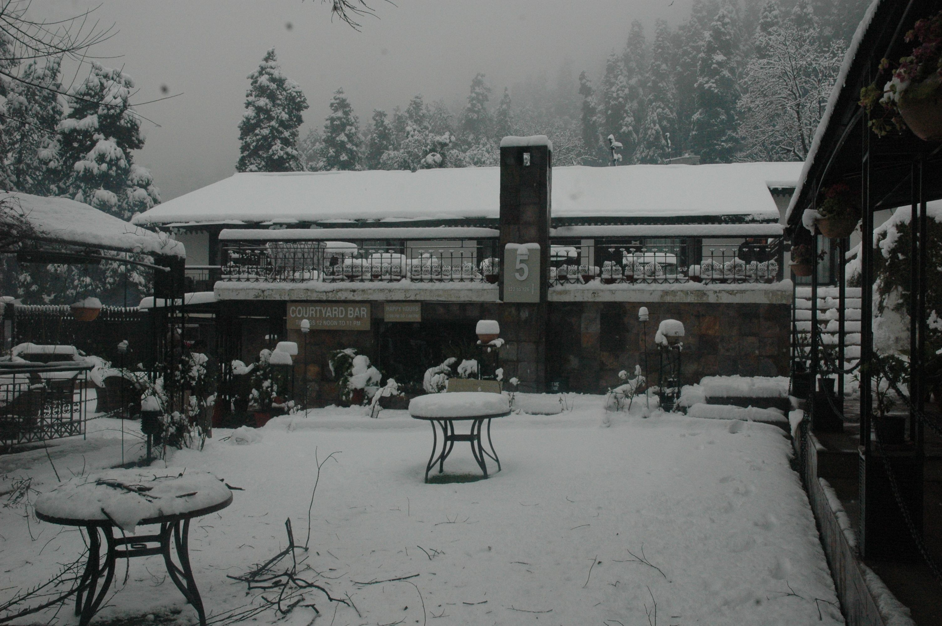 A courtyard with a snow-covered table and chairs, a sign for a courtyard bar, and a building with a snow-covered roof.