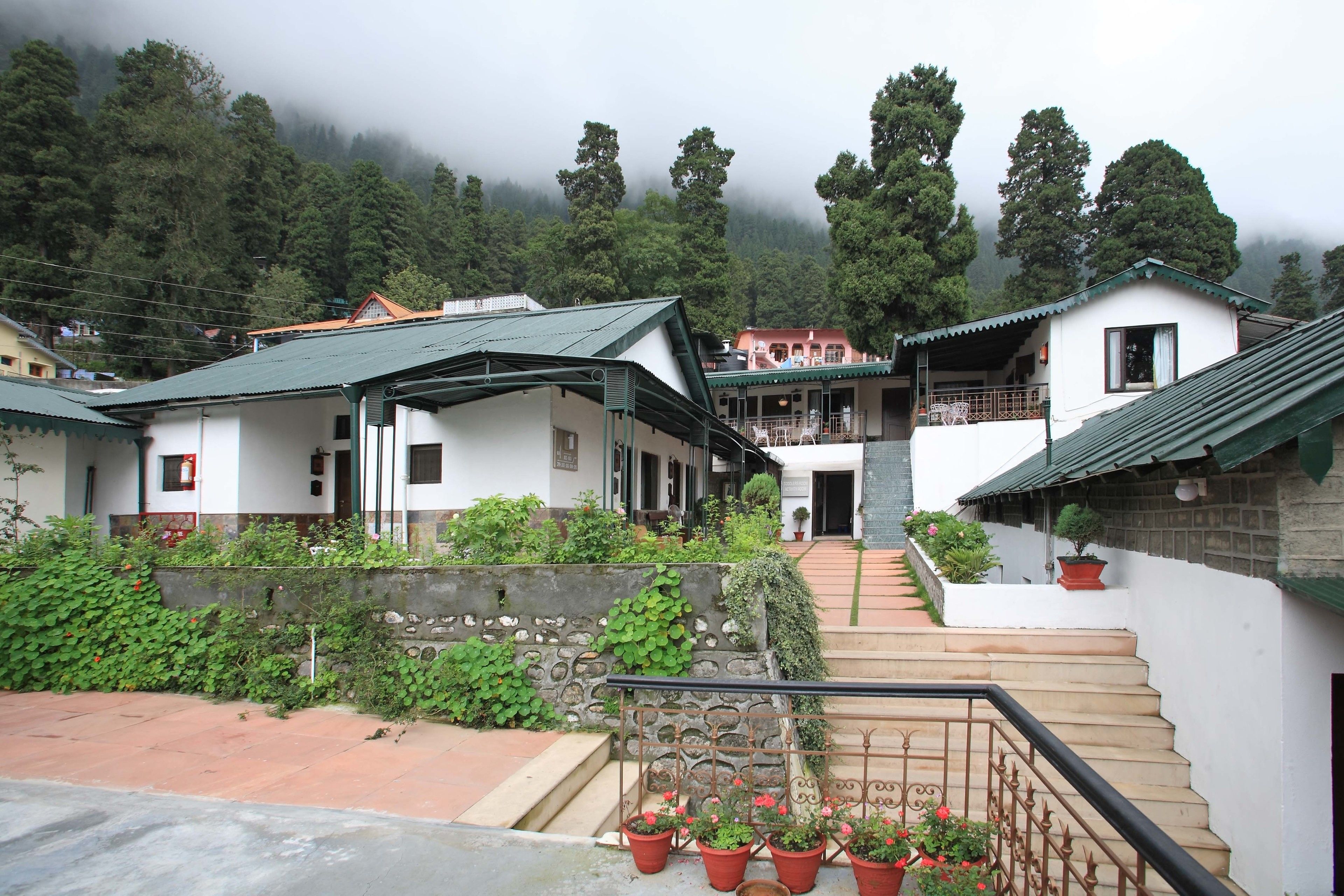 A traditional building with a staircase leading to a balcony, surrounded by lush greenery and a mountainous backdrop.