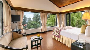 A hotel room with a bed, a sofa, a TV, a small table with a vase, and large windows with curtains.