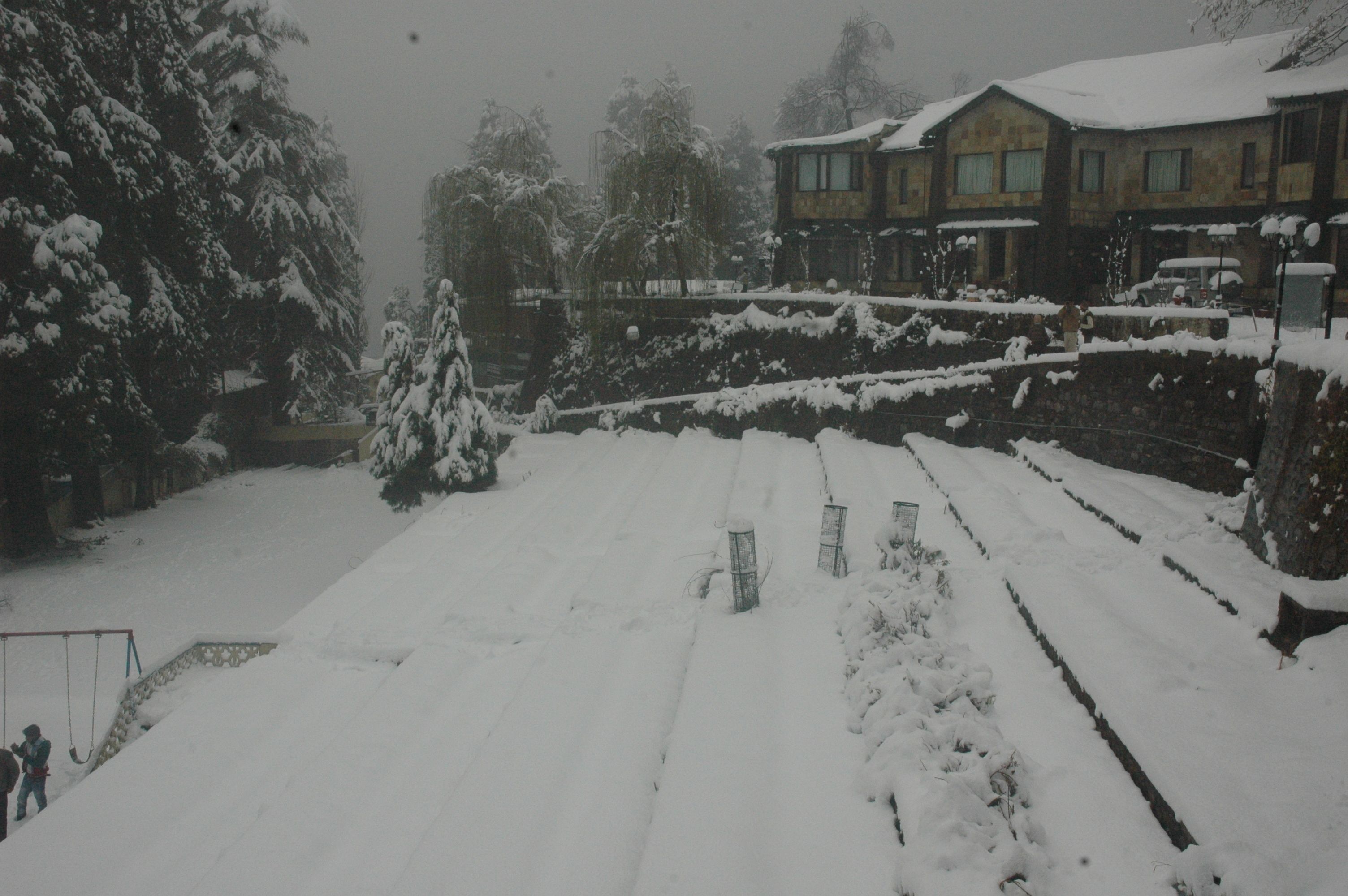 A snow-covered courtyard with a swing set, a stone wall, and a building with multiple windows.