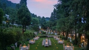 A garden venue with multiple round tables and chairs, arranged in rows, surrounded by lush greenery and trees.