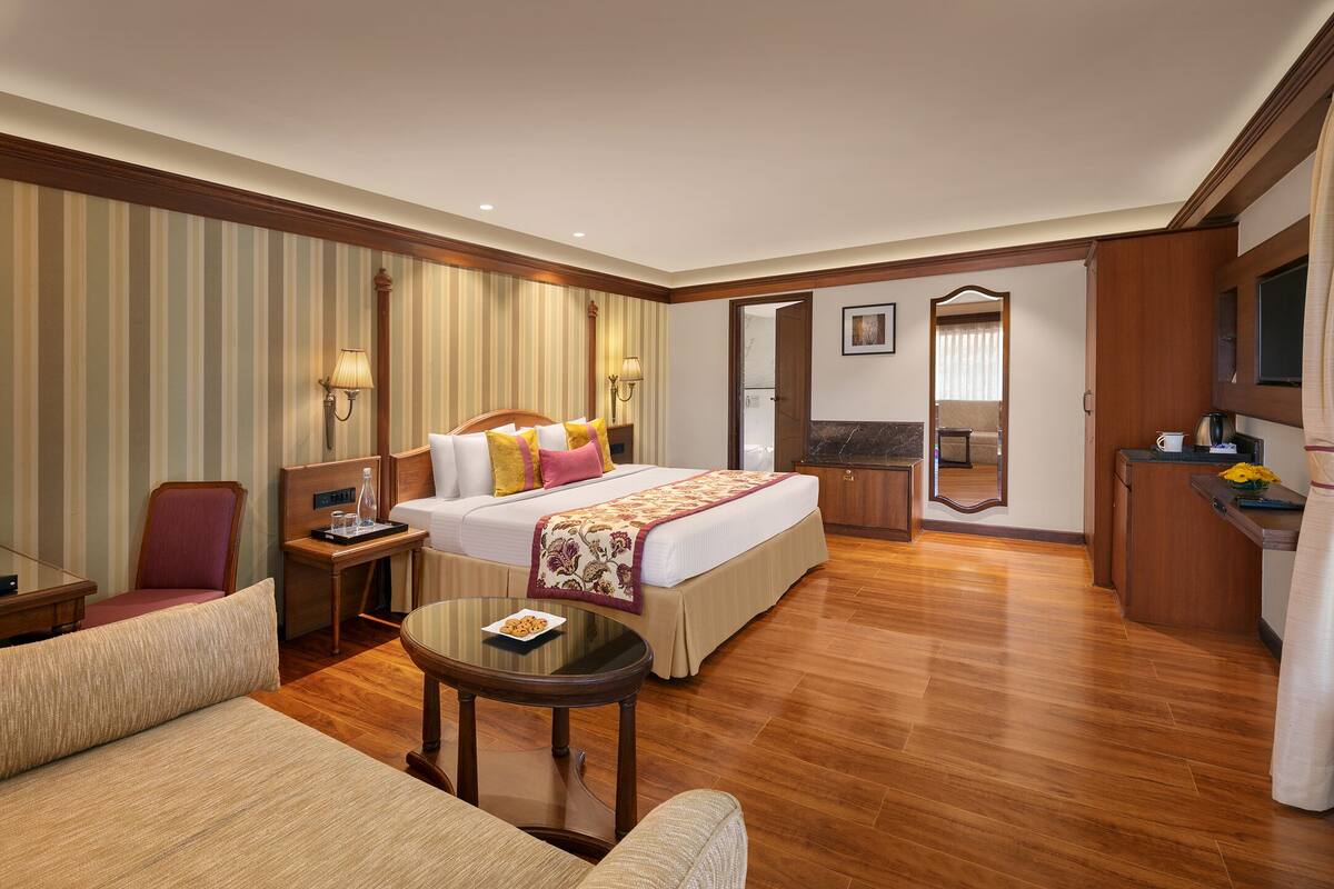 A hotel room with a large bed, a sitting area with a sofa and a small table, a desk with a chair, a mirror, and a television.