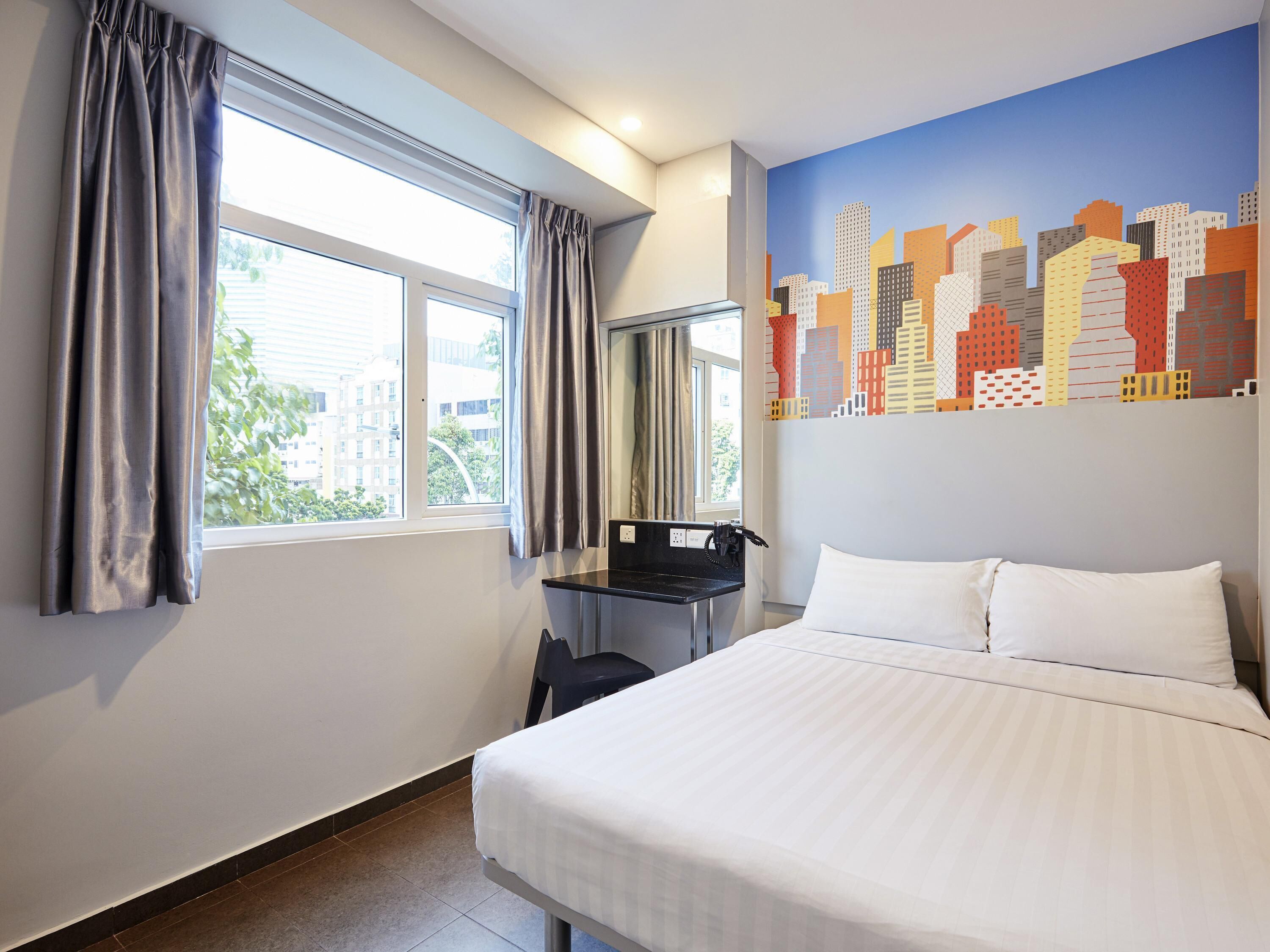 A hotel room with a bed, a desk with a chair, a window with curtains, and a cityscape mural on the wall.