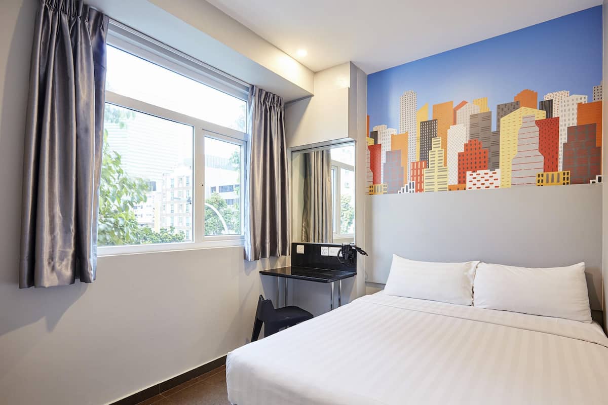 A hotel room with a bed, a desk with a chair, a window with curtains, and a cityscape mural on the wall.