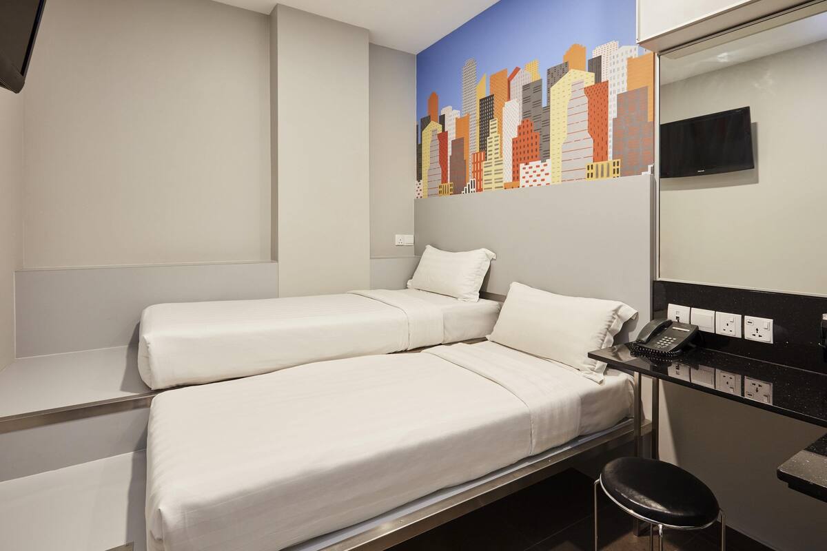 A hotel room with two beds, a desk with a telephone, and a cityscape mural on the wall.