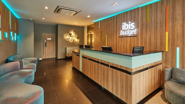 Un área de recepción moderna con un mostrador de madera, una zona de estar con sillones acolchados y un letrero que dice "ibis budget".