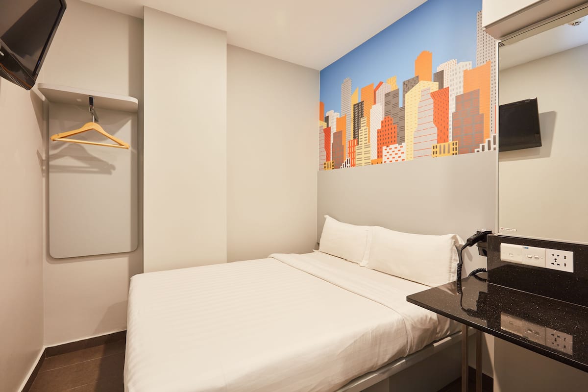 A hotel room with a bed, a hanging coat rack, a cityscape mural, a flat-screen TV, and a power outlet.