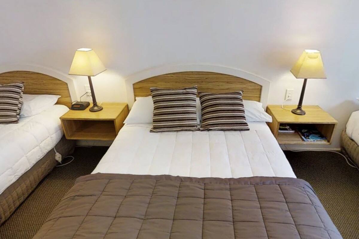 A hotel room with three single beds, each with a wooden headboard and bedside lamps.