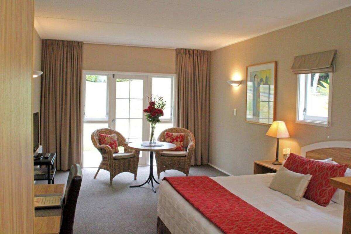 A hotel room with a bed, two chairs, a small table with a vase, a nightstand with a lamp, and a painting on the wall.