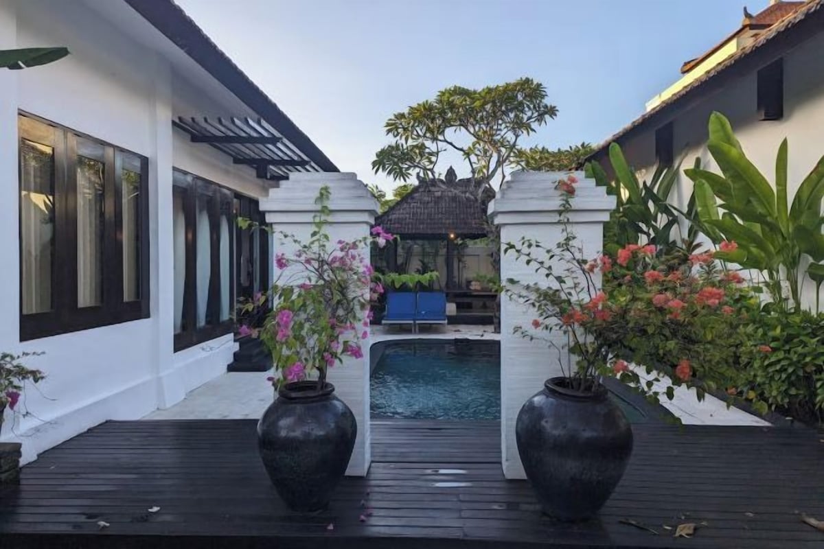 A pool area with a wooden deck, surrounded by white walls and tropical plants.