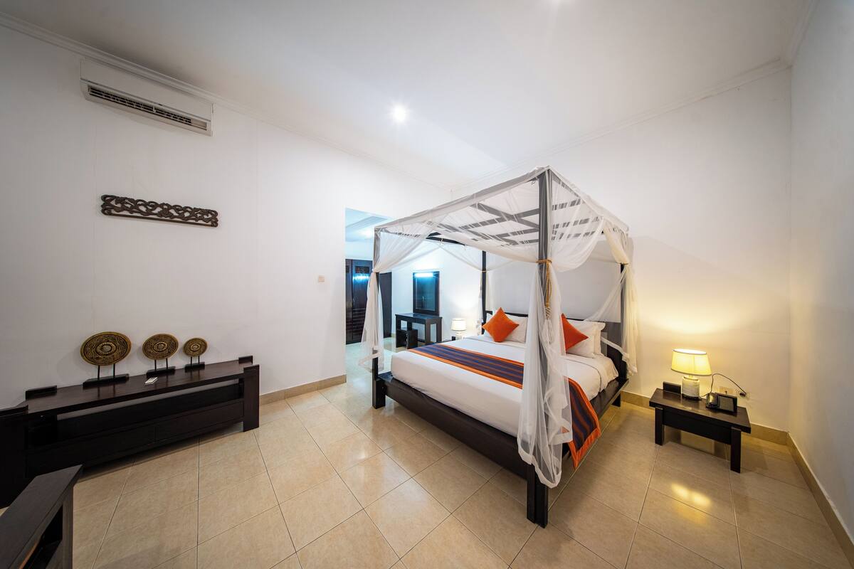 A modern hotel room with a four-poster bed, a bedside table with a lamp, and a decorative console table with two wall-mounted items.