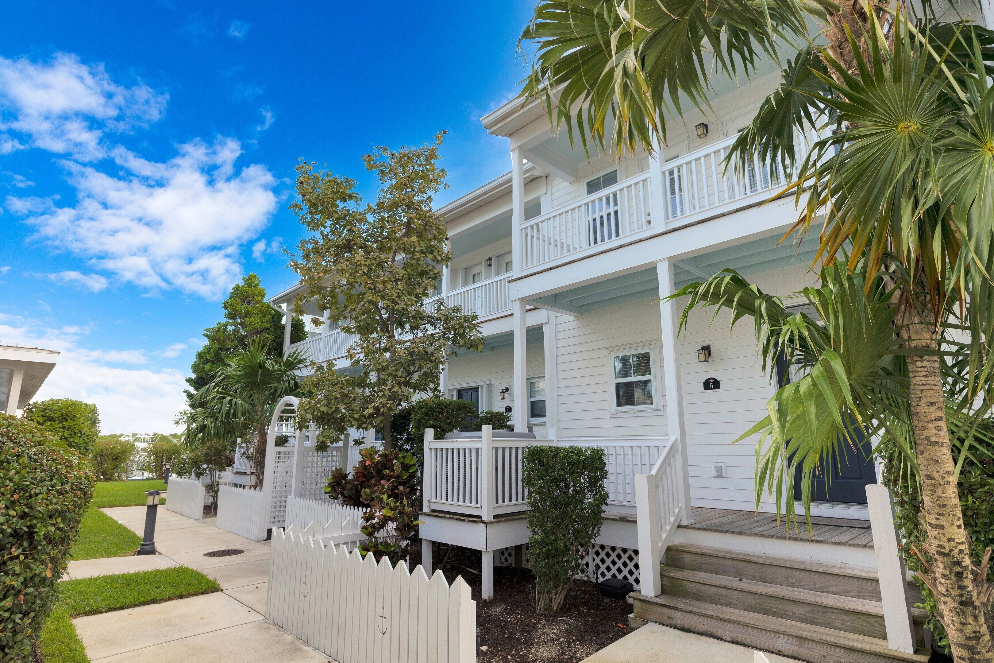 A two-story white building with a balcony, surrounded by greenery and a white picket fence.