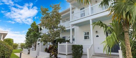 A two-story white building with a balcony, surrounded by greenery and a white picket fence.