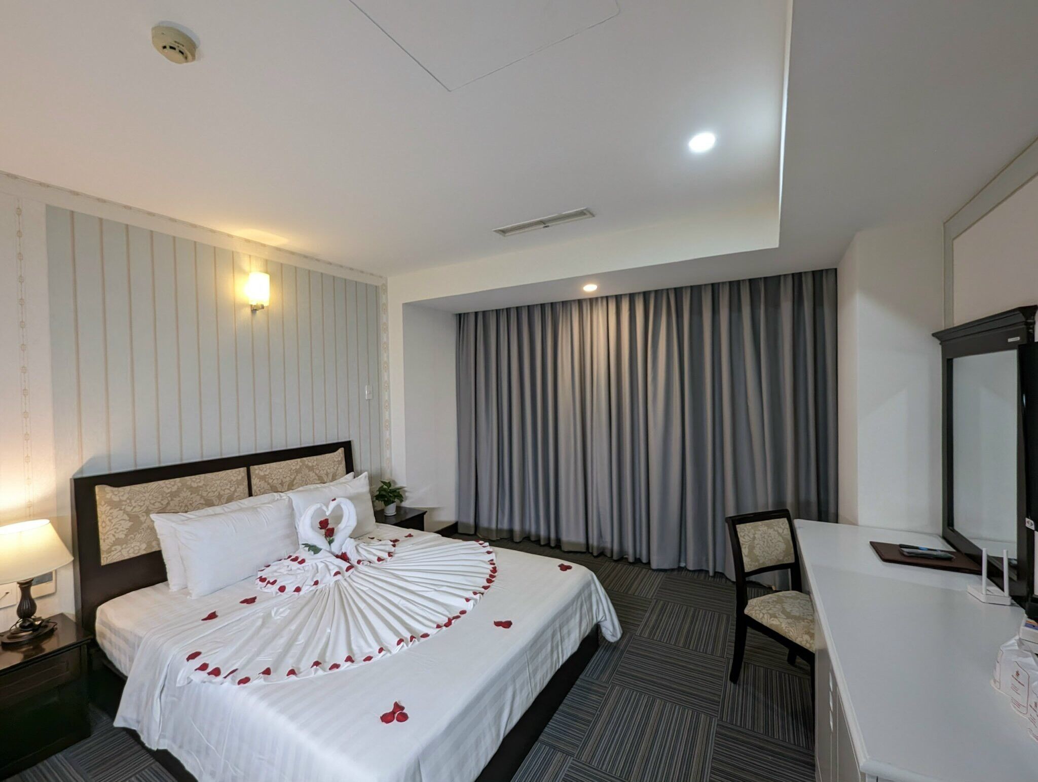 Une chambre d’hôtel avec un lit, un bureau, une chaise, une lampe et une grande fenêtre avec des rideaux.