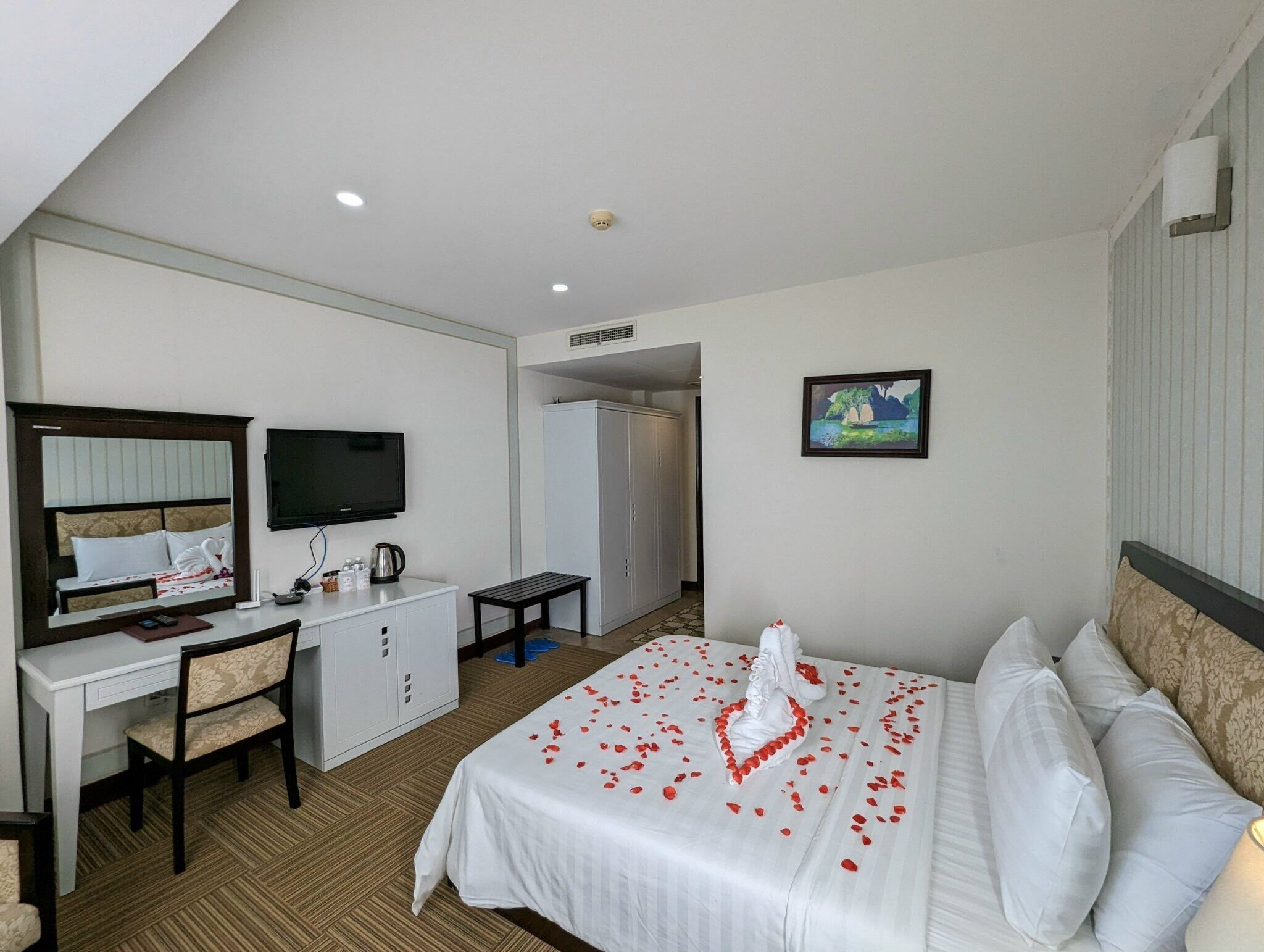 A hotel room with a bed, a desk, a chair, a television, a wardrobe, and a painting on the wall.