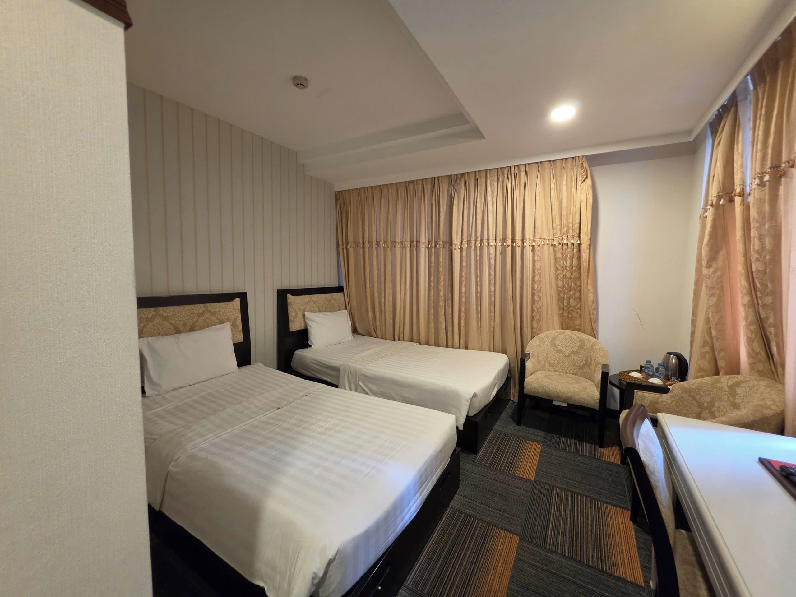 A hotel room with two beds, a chair, a small table with a lamp, and a window with curtains.
