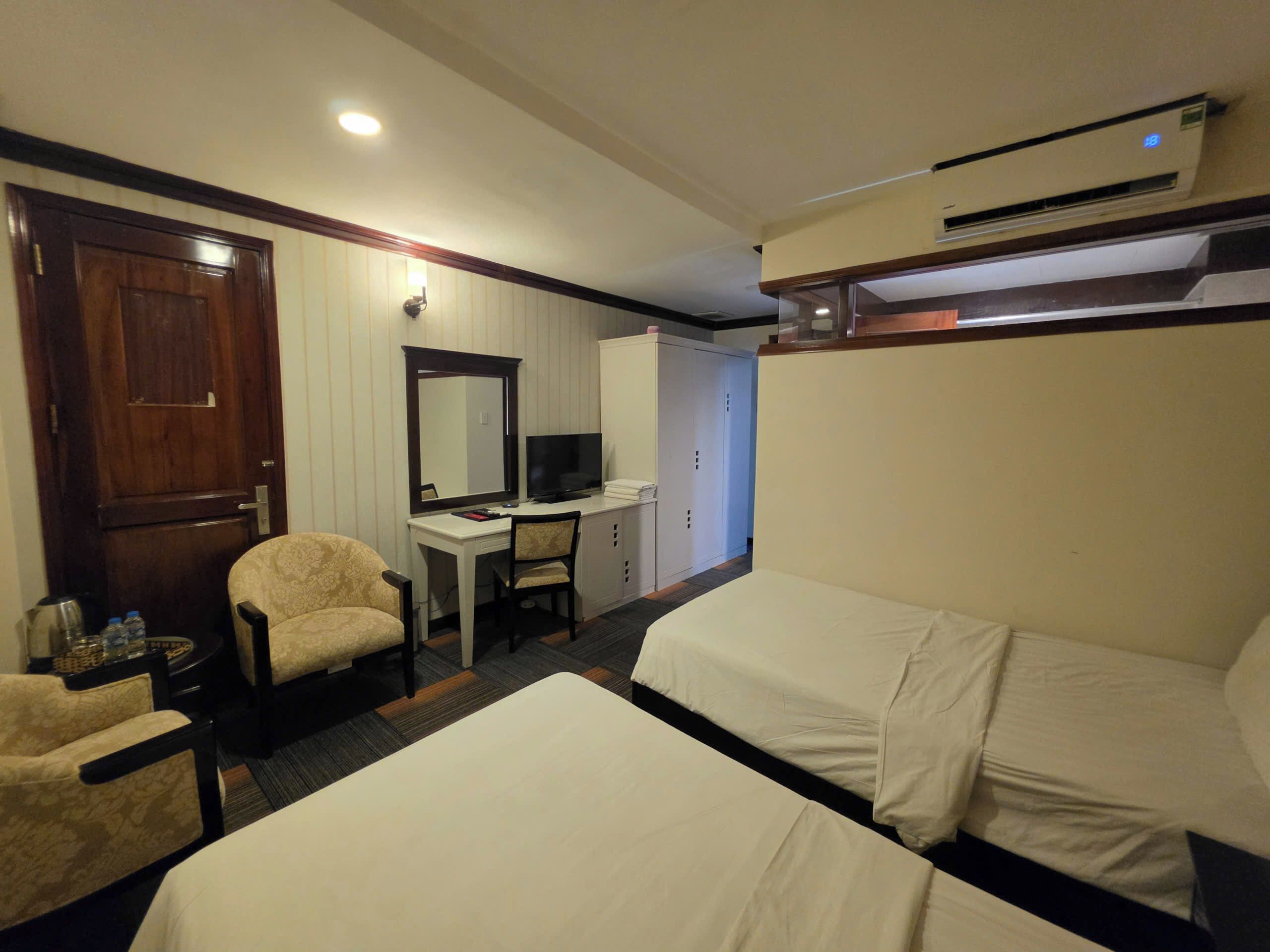 A hotel room with two beds, a desk with a television, a chair, a wardrobe, and an air conditioning unit.