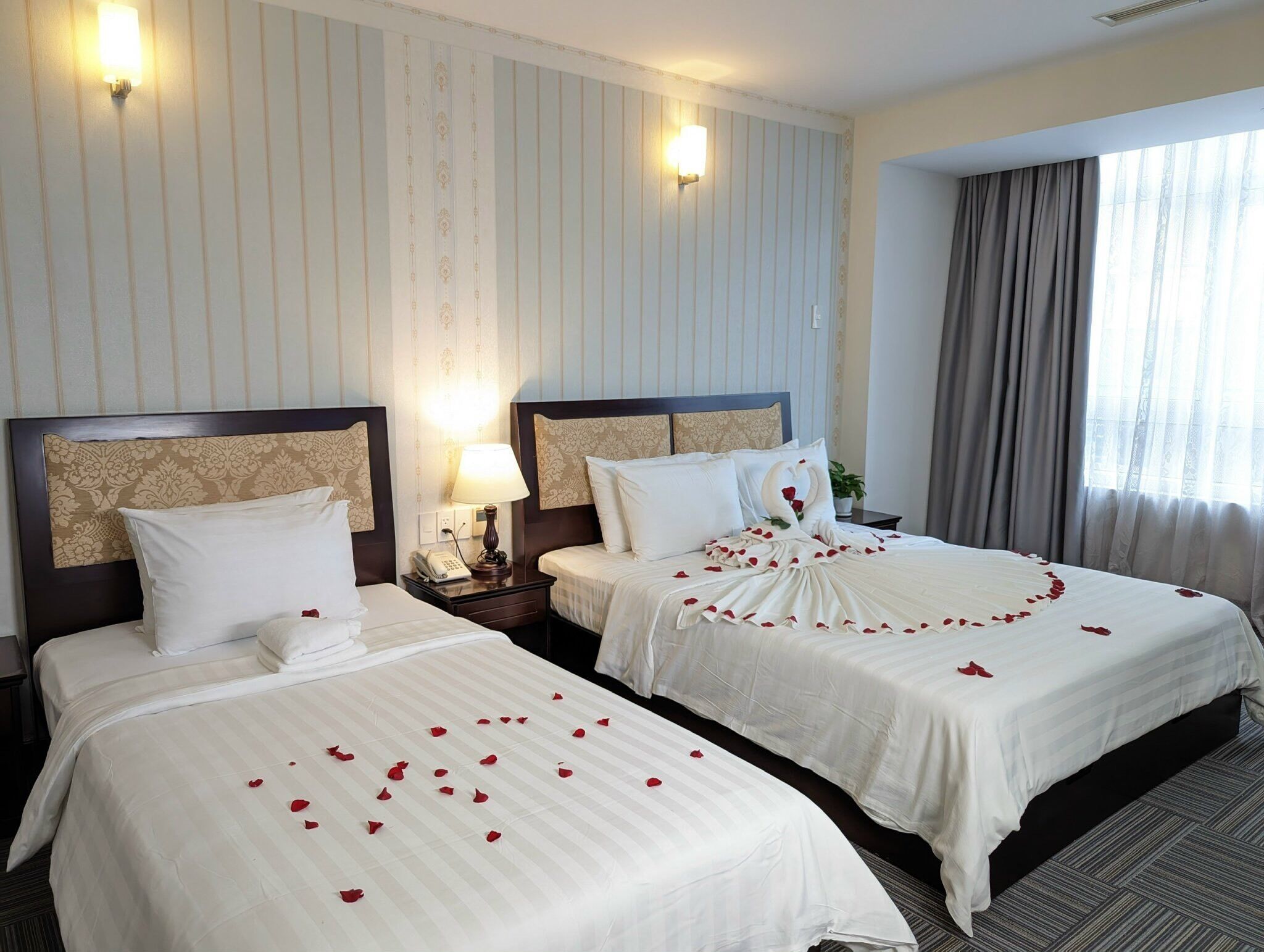 Une chambre d’hôtel avec deux lits, chacun agrémenté de draps blancs et de pétales de rose. On y trouve des lampes fixées au mur et une grande fenêtre avec des rideaux.