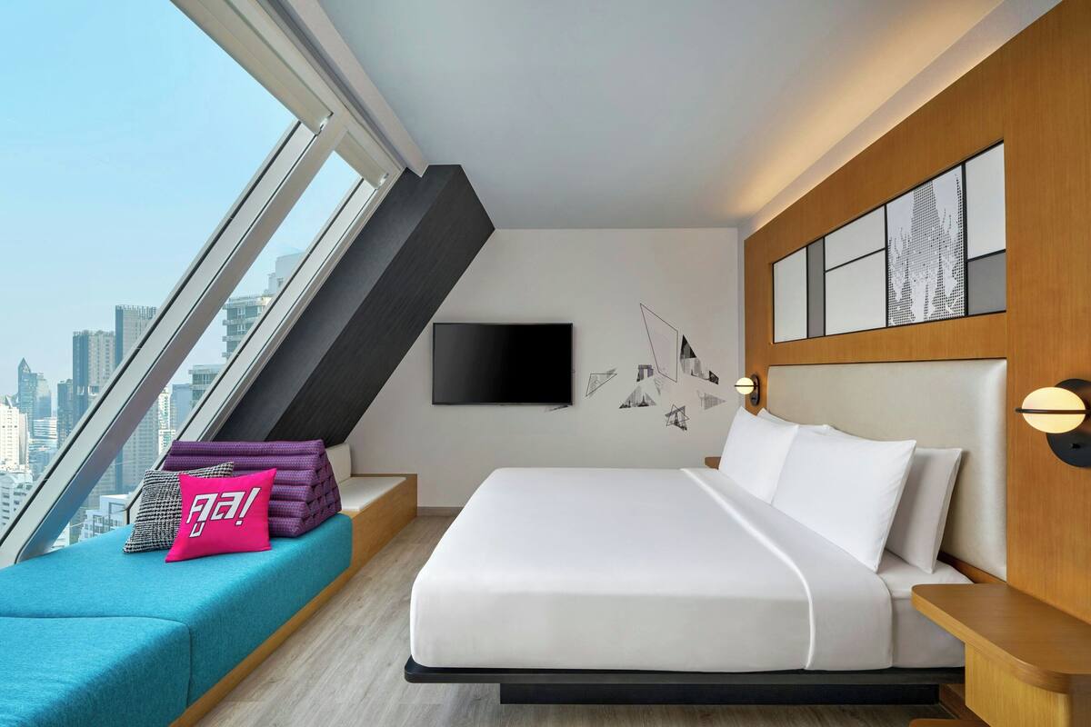 A modern hotel room with a large bed, a city view, a flat-screen TV, and a unique wall art installation.