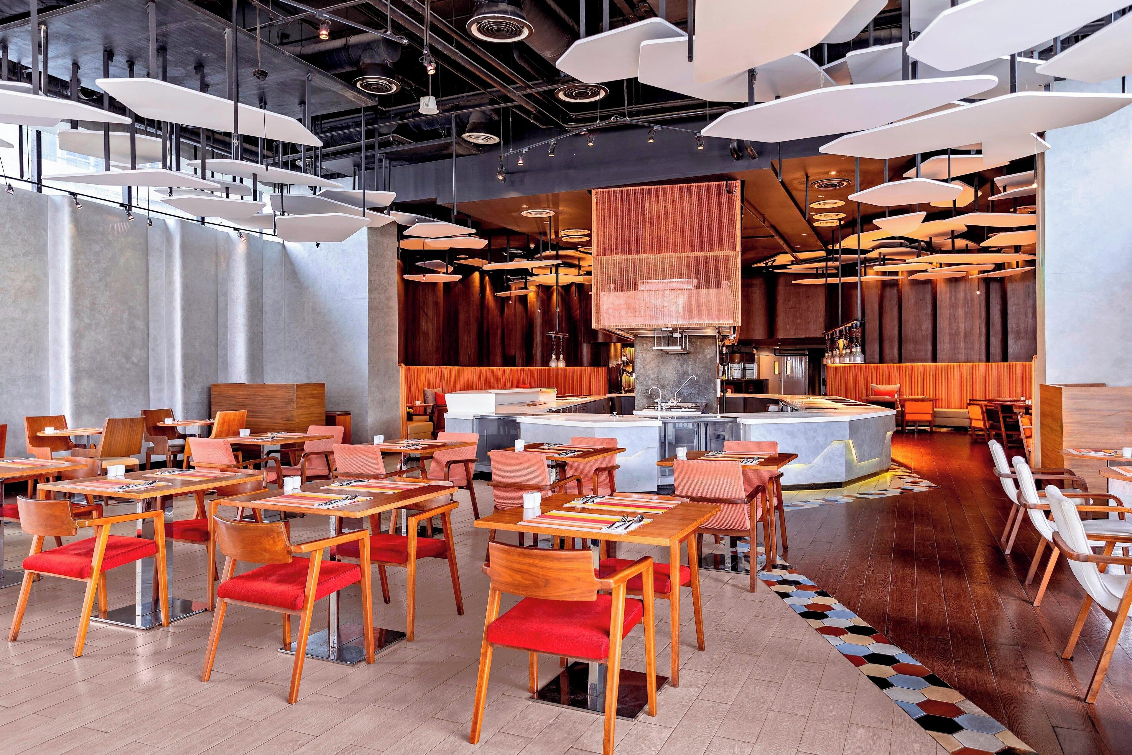 A modern restaurant with wooden tables and red cushioned chairs, a central bar area, and contemporary lighting fixtures.
