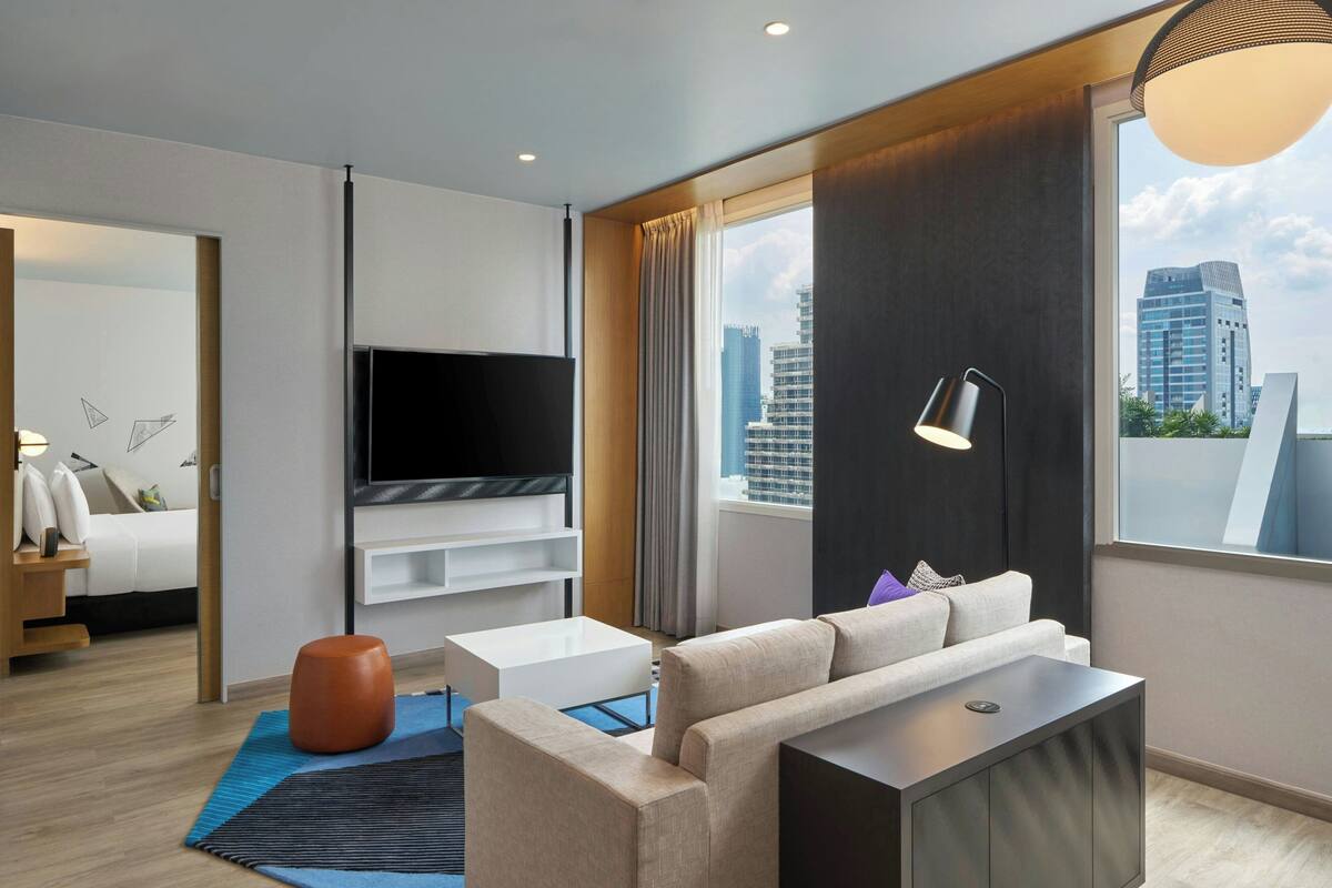 A modern hotel room with a large TV, a sofa, a coffee table, and a view of the city skyline.
